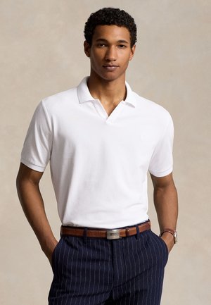 Νέος άνδρας που φοράει λευκό πόλο με κοντά μανίκια, μπουκαρισμένο σε μπλε παντελόνι με ρίγες και καφέ ζώνη, στέκεται με τα χέρια στις τσέπες.