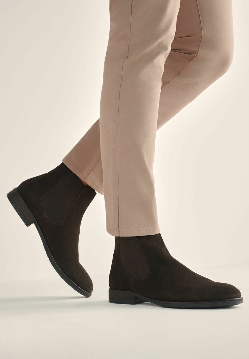 Brązowe zamszowe botki Chelsea z elastycznymi bocznymi panelami, okrągłym noskiem i niskim, warstwowym obcasem, w zestawieniu z jasnobeżowymi spodniami typu tapered.