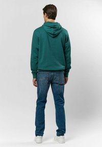 Teal kapucnis pulóver normál szabásban, kék farmerral párosítva. Részletezi a bordás mandzsettákat, és a hátulról készült képen a zsebek részletei láthatók.