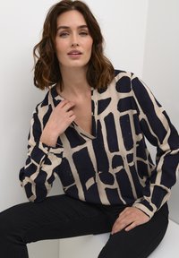Marineblauwe en beige gebloemde blouse met een V-hals en lange mouwen; gladde textuur met abstracte blokpatronen; gedragen met zwarte broeken.