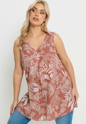 Kobieta o długich blond włosach, ubrana w bezrękawnik w kolorze rdzy z białym kwiatowym wzorem oraz niebieskie dżinsy, pozująca z rozluźnionymi ramionami.