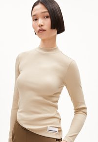 Beige geripptes Langarmoberteil mit Stehkragen; hat ein figurnahes Design und ein weißes Etikett mit "ARMEDANGELS" auf der unteren linken Seite.