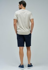 Camiseta de algodón beige de manga corta, combinada con shorts azul marino. El modelo se ve desde atrás, llevando zapatillas azules. Diseño simple y casual.