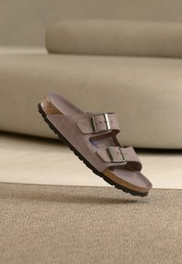 Ανοιχτό καφέ σαμουά σανδάλι με δύο ρυθμιζόμενες ασημένιες αγκράφες και πάτο από φελλό, τοποθετημένο σε μπεζ χαλί με φόντο έναν καναπέ.