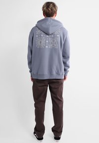 Sweat à capuche gris avec un cordon de serrage, présentant des motifs graphiques floraux et abstraits au dos. Porté avec un pantalon marron foncé et des baskets noires.
