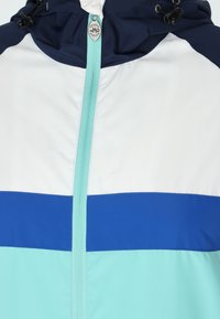 Farverig vindjakke med en marineblå hætte, hvid overdel og teal nederste sektioner. Central lynlås med turkis kant og logodetalje.