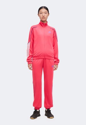 Person, der står iført en knaldrød Adidas træningsdragt med blå striber og sorte sneakers på en hvid baggrund.