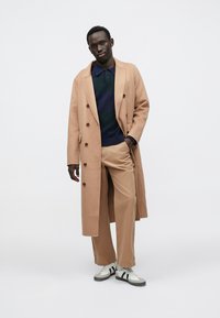 Manteau long beige clair avec boutons croisés, pull marine, pantalon beige et baskets blanches à rayures noires.