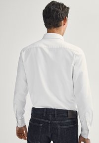 Camicia bianca a maniche lunghe con colletto classico e polsini a bottoni, indossata con jeans scuri. Il tessuto appare liscio e leggero.