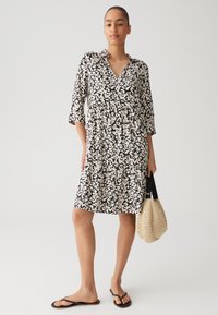 Black and white patterned dress with a v-neck, three-quarter sleeves, and tiered skirt. Paired with a woven beige bag and black flip-flops.