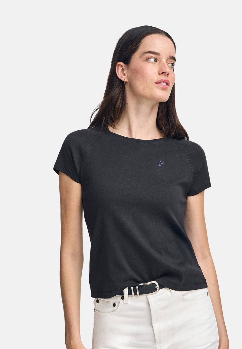 Svart kortärmad t-shirt i mjuk tyg med rund halsringning och en liten blå logotyp på bröstet, kombinerad med vita jeans och ett bälte.