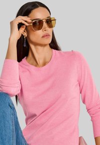 Rosafarbenes gestricktes Sweatshirt mit rundem Halsausschnitt und langen Ärmeln, kombiniert mit braun getönten Sonnenbrillen und hängenden lila Ohrringen.