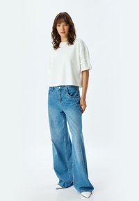 Witte crop top met textuuraccenten op de schouders, gecombineerd met lichtblauwe wijde jeans en puntige witte hakken, op een neutrale achtergrond.