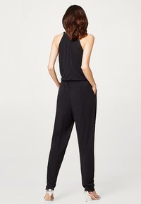 Black jumpsuit with a halter neckline, loose fit, and side pockets. Fabric appears smooth, lightweight, with tapered legs. Silver high heels worn.