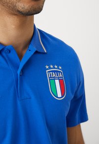 Μπλε πόλο μπλούζα με κολάρο με χρυσό περίγραμμα, φέρει σήμα της FIGC Ιταλίας με αστέρια και τριχρωμία ασπίδα. Υφασμάτινη υφή.