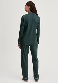 Dunkelgrünes, langärmliges Schlafanzug-Set mit weißer Paspelierung an den Bündchen und dem Kragen. Glattes Material, lockere Passform, gerade geschnittene Hosen.