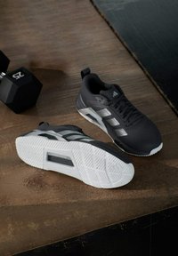 Chaussures de sport noires avec une tige texturée, des accents blancs et une semelle en caoutchouc à motifs, posées sur une surface en bois à côté d'un haltère.