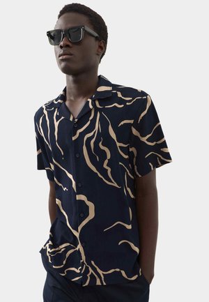 Młody mężczyzna w ciemnych okularach przeciwsłonecznych i czarnej koszuli z krótkim rękawem ozdobionej beżowymi abstrakcyjnymi falistymi liniami, z rękami w kieszeniach, zwrócony lekko w lewo.