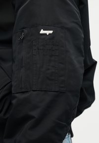 Μανίκι μαύρου μπουφάν με τσέπη που κλείνει με φερμουάρ και απλικέ ασημένιο λογότυπο «hugo» πάνω από ραμμένες ορθογώνιες λεπτομέρειες.