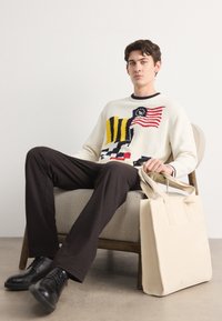 Pull en tricot avec une base crème, présentant un motif de drapeau en rouge, jaune et noir. Pantalons foncés, chaussures noires et un sac fourre-tout beige.