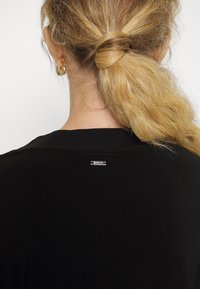 Schwarzes, leichtes Top mit einem gerippten Ausschnitt, das ein kleines silbernes Logo-Tag auf der Rückseite nahe dem Hals hat. Haare in lockeren Wellen frisiert.