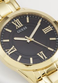 La montre-bracelet en ton or possède un cadran noir avec des chiffres romains. Le bracelet métallique a une finition brillante et un design en maillons, ajoutant de la durabilité.