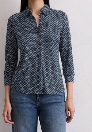 Femme portant une chemise ajustée bleu foncé à motifs géométriques blancs et un jean bleu, debout contre un mur blanc uni.