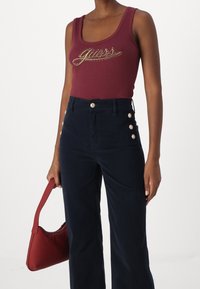 Top sin mangas burdeos con el logo dorado de "Guess", combinado con pantalones de tiro alto azul marino con detalles en dorado y un bolso rojo.