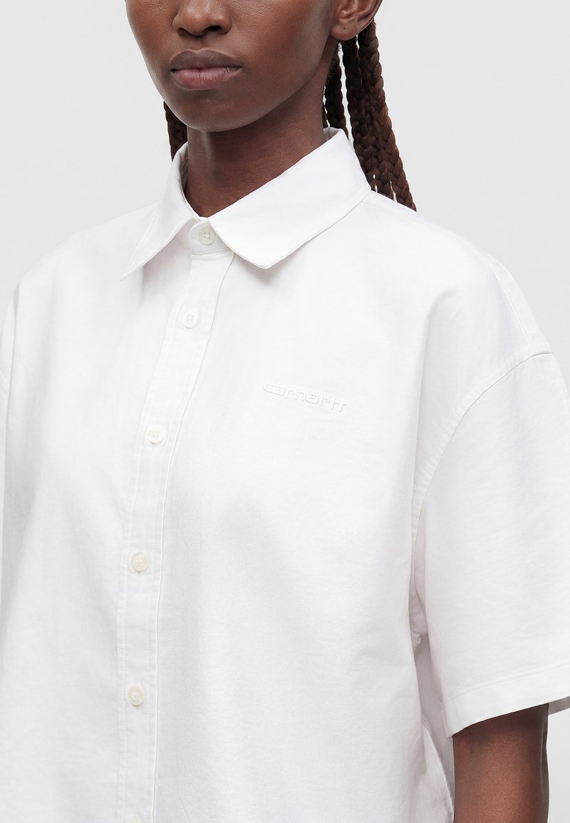 Person with braided hair wearing a white short-sleeve button-up shirt with a collar and subtle embroidered logo on the chest.