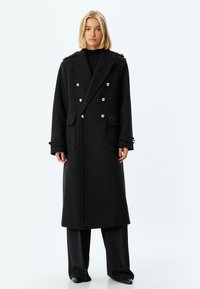 Abrigo negro de doble botonadura con botones dorados, bolsillos laterales y acentos en los hombros. Fabricado en una tela texturizada, con una silueta larga y recta.