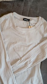 Top a maniche lunghe grigio chiaro con texture a coste, scollo rotondo e etichetta attaccata; presenta un tessuto liscio e un design aderente.