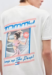 Πίσω μέρος λευκού μπλουζακιού με ρετρό γραφικό μιας γυναίκας σε ροζ ρούχα γυμναστικής που τρέχει, κείμενο "Tommy," "85,'" και "Jump up the Prep!"