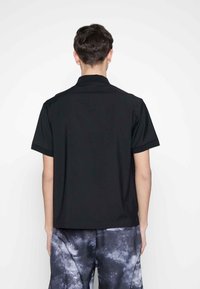 Černé tričko s krátkým rukávem, uvolněného střihu, plochým límečkem, hladkou texturou a bez viditelných vzorů nebo akcentů, zobrazené ze zadní strany.