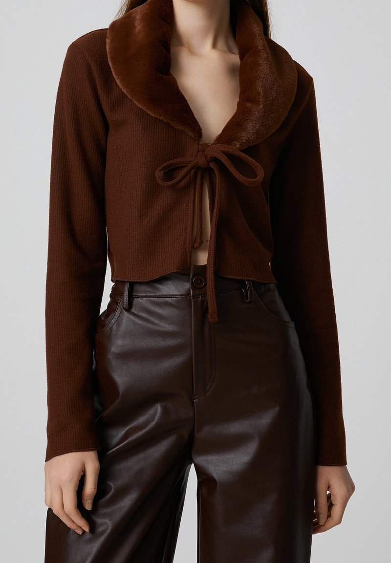 Femme portant un pull court marron à manches longues avec un col en fourrure noué à l'avant, associé à un pantalon en cuir marron foncé taille haute.