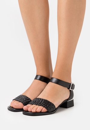 Sorte lædersandaler med en vævet rem og ankelspænde, med en lav blokhæl og åbent tådesign. Glat tekstur og minimale detaljer.