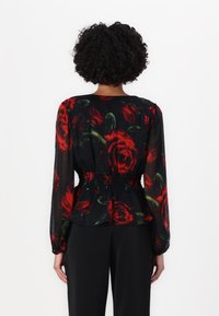 Blusa a maniche lunghe in tessuto trasparente nero con motivo di rose rosse, vita arricciata e vestibilità ampia. Abbinata a pantaloni neri.