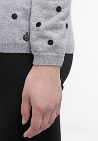 Main reposant sur le côté, vêtue d'un pull gris à pois porté sur un pantalon noir, sur un fond uni.