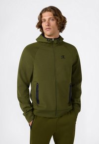 Jonge man met golvend haar, gekleed in een olijfgroene hoodie met ritssluiting en bijpassende broek, met zijn handen in zijn zakken, op een effen witte achtergrond.