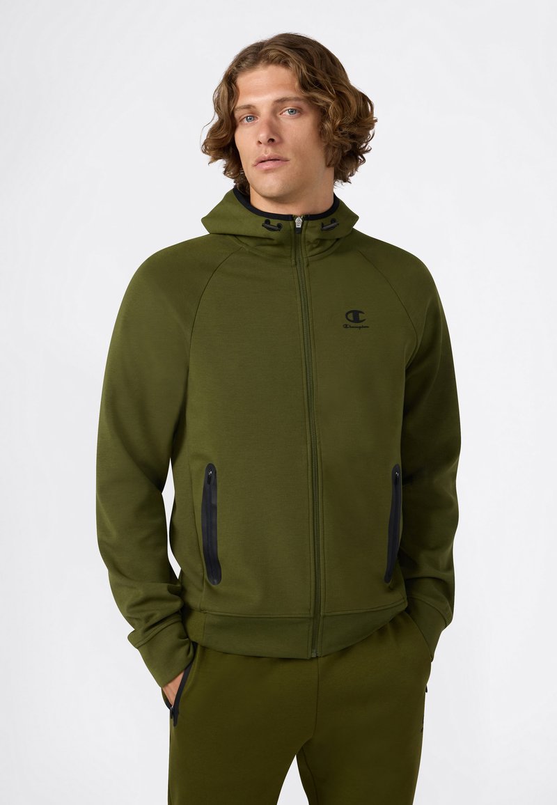 Jonge man met golvend haar, gekleed in een olijfgroene hoodie met ritssluiting en bijpassende broek, met zijn handen in zijn zakken, op een effen witte achtergrond.
