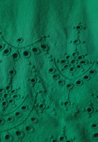 Tissu vert présentant une broderie complexe avec des découpes circulaires et des motifs floraux, mettant en valeur une surface texturée.