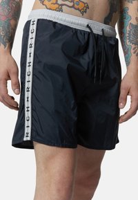 Pantaloni da bagno navy con cintura elastica, cordoncino e striscia laterale bianca a contrasto con la scritta "RICH" ripetuta. Tessuto leggero.