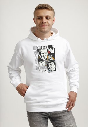 Junger Mann trägt einen weißen Star Trek Hoodie mit grafischen Comic-Porträts und Text, steht mit den Händen in den Taschen und lächelt leicht.