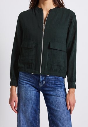 Frau trägt eine dunkelgrüne Reißverschlussjacke mit zwei vorderen Klappentaschen und blaue Jeans vor einem schlichten Hintergrund.