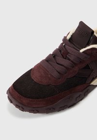 Μπορντό και μαύρο αθλητικό παπούτσι με σουέντ και υφή υλικού, στρογγυλά κορδόνια, λαστιχένια σόλα με μοτίβο, και λογότυπο στη γλώσσα του παπουτσιού.