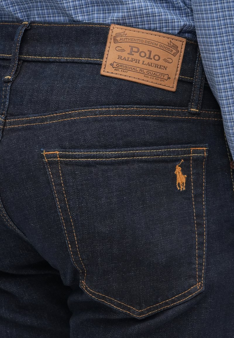 Mørkeblå denimjeans med orange syninger, der har et lædermærke og et lille broderet hestelogo på baglommen.