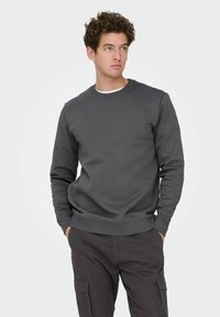 Jeune homme aux cheveux bouclés portant un sweat-shirt gris foncé et un pantalon cargo, debout les mains dans les poches, regardant vers la droite.