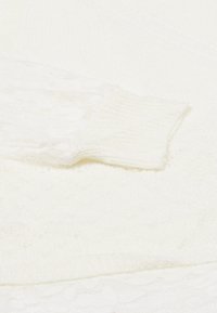 Lähikuva valkoisesta neulotusta vaatteesta, jossa on ribbejä hihassa ja pitsiyksityiskohtia pehmeän teksturoidun kankaan taustalla.