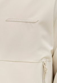 Jasnobeżowy materiał kurtki z małym logo Jack & Jones oraz pasującym beżowym zamkiem błyskawicznym z pętelką po prawej stronie na dole.