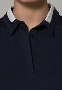 Laivastonsininen poolopaita, jossa on valkoinen kaulus, kolmella napilla varustettu napitus ja resorikuvioinen kangas. Selkeät sauman yksityiskohdat näkyvissä.