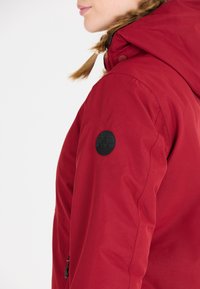 Röd vattentät jacka med huva, dragkedjefickor och en svart cirkulär logotyp på ärmen. Texturerad tyg med synliga sömmar.